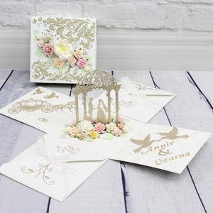 Op de afbeelding: Een witte vierkante pop-up kaart met gouden glitterfiligraan, een centrale prieelstructuur met een silhouet van een koppel en kleine papieren bloemen. De kaartpanelen tonen gouden glitterillustraties van een koets, vogels en de tekst Annie & Oeasna.