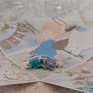 Könnte beinhalten: Eine mehrlagige Explosionsbox-Karte aus Papier mit einem mittig platzierten, gestanzten Engel mit glitzerndem Heiligenschein und Flügeln. Die Karte ist mit blau-weißen Blumenmustern, glitzernden goldenen Wimpeln und dreidimensionalen Papierrosen verziert.