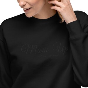 Może przedstawiać: Czarna bluza z napisem "Mom Up" w kursywie. Poniżej tekstu znajduje się fraza "life is not mini". Bluza jest w jednolitym czarnym kolorze.