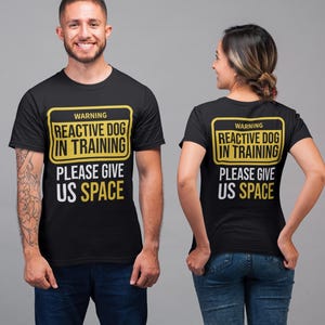 May include: Black t-shirts with a yellow and white warning label that reads "WARNING REACTIVE DOG IN TRAINING PLEASE GIVE US SPACE." The shirts are worn by two people. The background is a solid gray.
