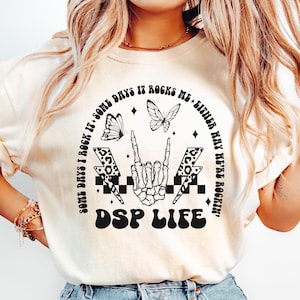May include: Cream-colored t-shirt with a graphic design featuring butterflies, lightning bolts, and a skeleton hand making a rock-and-roll gesture. The text reads "Some days I rock it. Some days it rocks me. Either way we're rockin'. DSP LIFE."