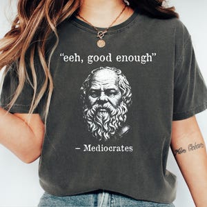 May include: Dark grey heather t-shirt with a white graphic of a philosopher and the text "eeh, good enough" above the image and "- Mediocrates" below. The shirt has a crew neck.