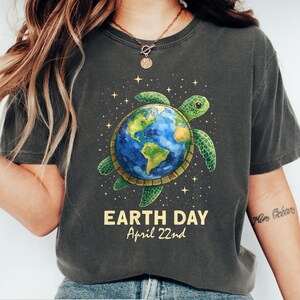 May include: Dark grey heather t-shirt featuring a green sea turtle carrying the Earth. The Earth is depicted in blue and green, surrounded by stars. The text "EARTH DAY April 22nd" is printed below the turtle. The shirt is a casual wear item.