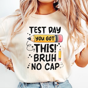 May include: Cream-colored t-shirt with the text "TEST DAY YOU GOT THIS! BRUH NO CAP" in black, yellow, and pink. The design includes a large pencil graphic and stars. The shirt is made of a soft material.