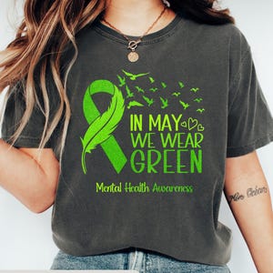 Puede incluir: Camiseta gris oscuro jaspeado con una cinta verde y el texto "IN MAY WE WEAR GREEN" y "Mental Health Awareness". El dise&ntilde;o incluye una pluma y p&aacute;jaros. Un estilo casual y c&oacute;modo.
