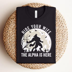 Puede incluir: Camiseta negra con un gráfico blanco de un hombre lobo aullando a la luna llena. El texto "HIDE YOUR WIFE THE ALPHA IS HERE" está impreso encima y debajo del gráfico. La camiseta está doblada sobre una superficie tejida.
