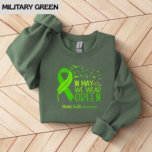 Puede incluir: Sudadera verde militar con un dise&ntilde;o de cinta y pluma verde brillante. El texto dice "IN MAY WE WEAR GREEN" y "Mental Health Awareness". Las mangas est&aacute;n enrolladas. La sudadera est&aacute; sobre una superficie de madera.
