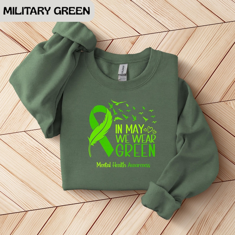 Puede incluir: Sudadera verde militar con un dise&ntilde;o de cinta y pluma verde brillante. El texto dice "IN MAY WE WEAR GREEN" y "Mental Health Awareness". Las mangas est&aacute;n enrolladas. La sudadera est&aacute; sobre una superficie de madera.