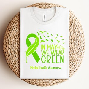 Puede incluir: Camiseta blanca con un dise&ntilde;o de cinta y pluma verdes, acompa&ntilde;ada del texto "IN MAY WE WEAR GREEN" y "Mental Health Awareness". El dise&ntilde;o incluye p&aacute;jaros verdes. La camiseta est&aacute; doblada sobre una superficie tejida.