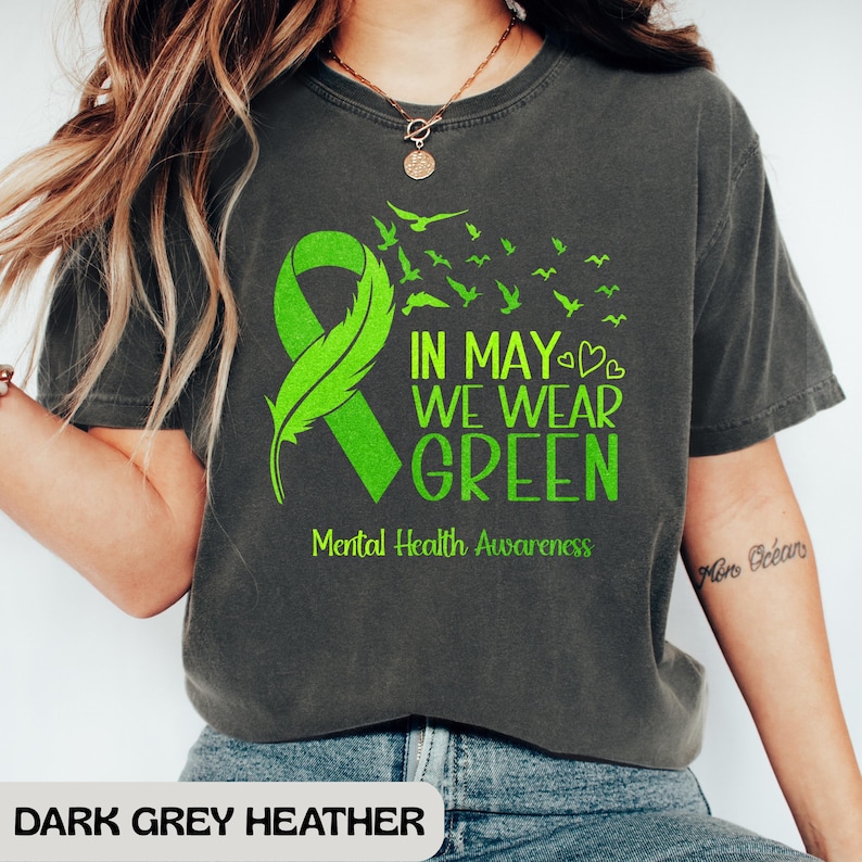 Puede incluir: Camiseta gris oscuro jaspeado con una cinta verde y el texto "IN MAY WE WEAR GREEN" y "Mental Health Awareness". El dise&ntilde;o incluye una pluma y p&aacute;jaros. Un estilo casual y c&oacute;modo.