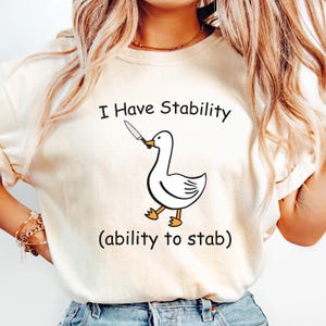 May include: Cream-colored sweatshirt with the text "I Have Stability" above a cartoon duck holding a knife. Below the duck, the text reads "(ability to stab)". The duck is white with black outlines and orange feet.