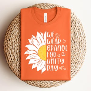 May include: An orange t-shirt featuring a white graphic of a half sunflower on the left side. The right side displays the white text "WE WEAR ORANGE FOR UNITY DAY" accompanied by small hand-drawn heart and flower icons in a clean, sans-serif font.