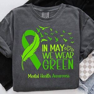 Puede incluir: Sudadera gris oscuro con un dise&ntilde;o de cinta y pluma verdes. El texto "IN MAY WE WEAR GREEN" y "Mental Health Awareness" est&aacute;n impresos en verde. Tambi&eacute;n presenta una bandada de p&aacute;jaros verdes.