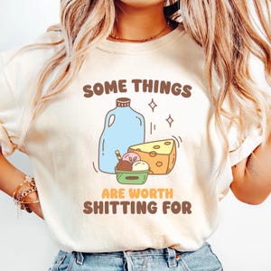 May include: Cream-colored sweatshirt with the phrase "SOME THINGS ARE WORTH SHITTING FOR" in brown lettering. The graphic includes a blue milk jug, a wedge of cheese, and a bowl of ice cream.