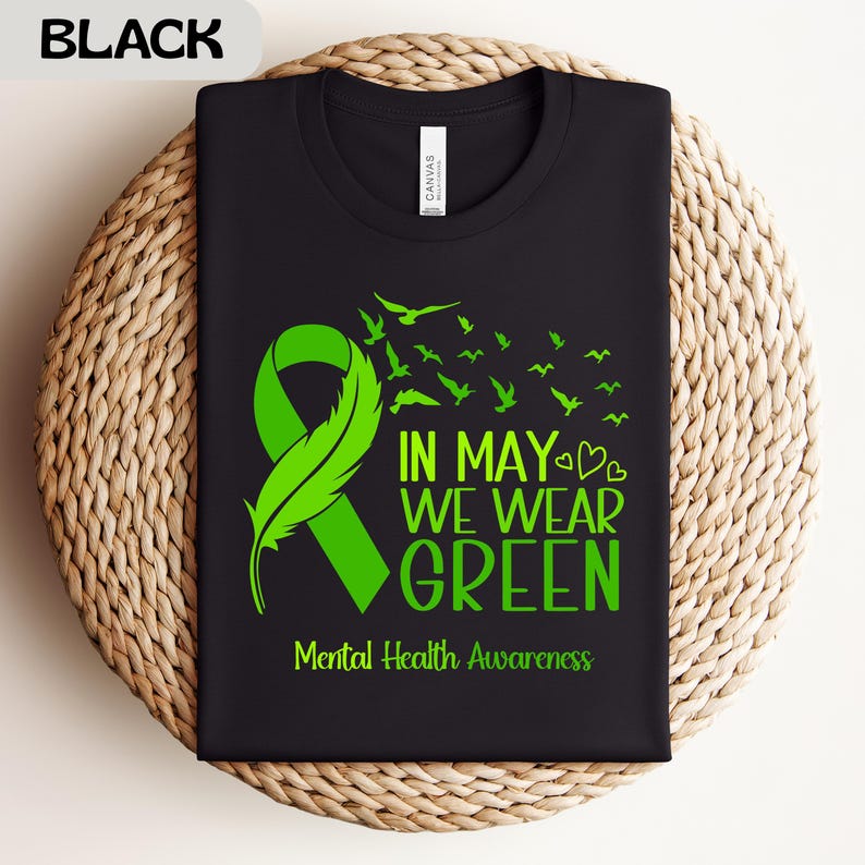 Puede incluir: Camiseta negra con un dise&ntilde;o de lazo y pluma verdes, acompa&ntilde;ada del texto "IN MAY WE WEAR GREEN" y "Mental Health Awareness". El dise&ntilde;o incluye p&aacute;jaros volando. La camiseta se muestra sobre una superficie tejida.