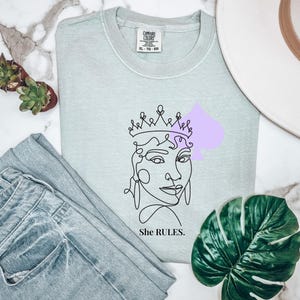May include: A light green crew neck sweatshirt with a line drawing of a woman wearing a crown and a purple spade shape. The text "She RULES." is below the drawing. The sweatshirt is paired with light blue jeans.