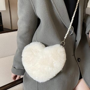 May include: A fluffy, heart-shaped, off-white purse with a pearl strap. The purse is made of a soft, textured material. The strap is a string of small, white pearls. The purse is worn over a gray blazer.