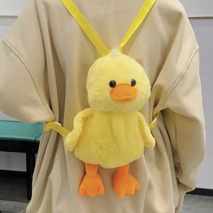 May include: A yellow plush duck backpack with adjustable yellow straps. The duck has a fluffy body, orange beak and feet, and black eyes. The backpack is worn over the shoulders.