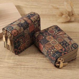 Puede incluir: Dos carteras rectangulares de corcho con un diseño de patchwork floral en tonos azul marino, marrón y naranja. Cada cartera tiene un cierre de cremallera dorado. Las carteras se exhiben sobre una superficie de madera.
