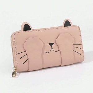 May include: A pale pink wallet with a cat face design. The wallet has a zip closure and a gold-coloured zip pull. The cat design includes black whiskers, a nose, and ears with black tips. The wallet is rectangular in shape.