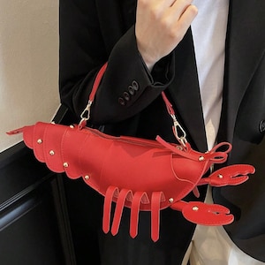 Op de afbeelding: Een rode handtas in de vorm van een kreeft met goudkleurige klinknagels en een ritssluiting. De tas heeft een lange, slanke vorm met klauwachtige details en een schouderriem. De tas is gemaakt van een glad materiaal.