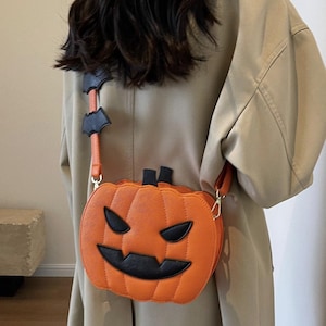 May include: Orange pumpkin-shaped crossbody bag with black facial features and a black stem. The strap is orange with black bat-shaped accents. A fun accessory for Halloween.