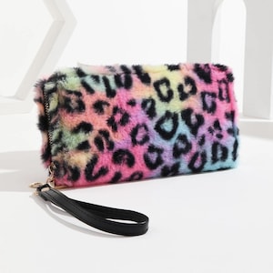 Puede incluir: Un bolso de mano rectangular con estampado de leopardo arcoíris. La bolsa de piel sintética presenta manchas negras sobre una mezcla de rosa, amarillo, azul y verde. Tiene una correa de muñeca negra y una cremallera dorada.