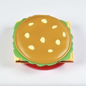 May include: A novelty wallet in the shape of a hamburger. It has a brown bun with white sesame seeds, green lettuce, yellow cheese, and a red tomato base. The wallet is designed to look like a classic burger.
