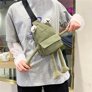 Peut inclure: Sac bandoulière en forme de grenouille vert clair avec une sangle noire. Le sac présente des yeux blancs avec des croix noires, une petite poche avant et de longues pattes pendantes. Le sac est porté en bandoulière.
