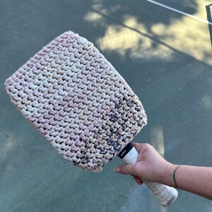 May include: A hand-crocheted tennis racket cover in a mix of cream, pink, and gray yarns. The cover is rectangular and fits over the racket head. The racket handle is white with black text.