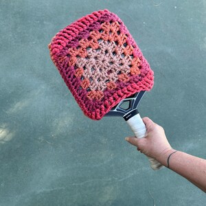 May include: A pickleball paddle with a handmade crochet cover. The cover features a granny square design in shades of pink, orange, and peach. The paddle has a black frame and a white grip.