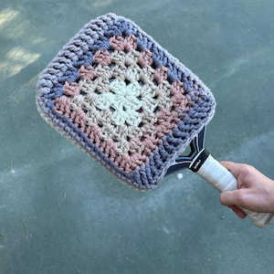 Puede incluir: Una funda de raqueta de tenis de ganchillo con un patrón de granny square. La funda presenta un diseño cuadrado con un cuadrado central crema, rodeado de cuadrados rosas, beiges y azul grisáceo. El mango de la raqueta es blanco.