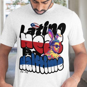 Op de afbeelding: Lichtgrijs t-shirt met een graffiti-stijl ontwerp met de tekst "Latino Heat Halftime". Het ontwerp bevat een voetbal met de Puerto Ricaanse vlag, een persoon met een zonnebril en konijnenoren en de tekst "Est. 2026."