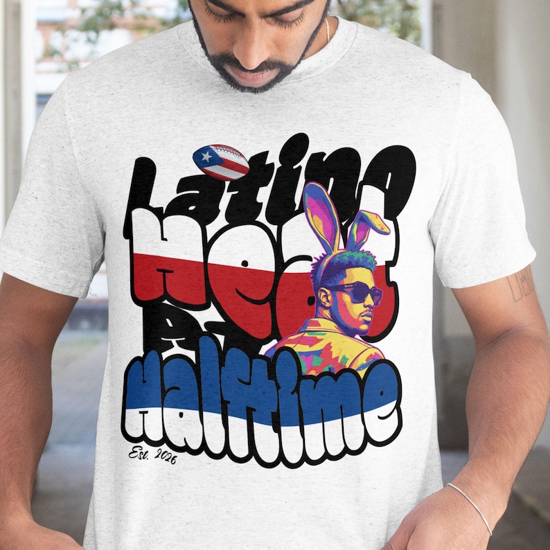 Op de afbeelding: Lichtgrijs t-shirt met een graffiti-stijl ontwerp met de tekst "Latino Heat Halftime". Het ontwerp bevat een voetbal met de Puerto Ricaanse vlag, een persoon met een zonnebril en konijnenoren en de tekst "Est. 2026."