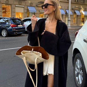 Könnte beinhalten: Eine lange, offene schwarze Strickjacke über einem weißen Kleid, kombiniert mit einer cremefarbenen Ledertasche mit braunem Innenfutter, Kordelzug und dünnen Schulterriemen, ergänzt durch eine schwarze Sonnenbrille und goldene Creolen.