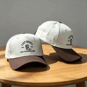 Może przedstawiać: Dwie czapki bejsbolowe z białymi koronami i brązowymi daszkami leżą na drewnianym stole. Czapki mają napis "LAST RODEO" i "LAKE WINNIPESAUKEE" z grafiką buta. Czapki są casualowe i odpowiednie do noszenia na zewnątrz.