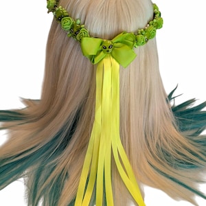 May include: A green floral crown with small green roses and alien head accents. A bright green bow with an alien head is centered on the crown, with long yellow ribbons cascading down. The hair is blonde with teal and green streaks.