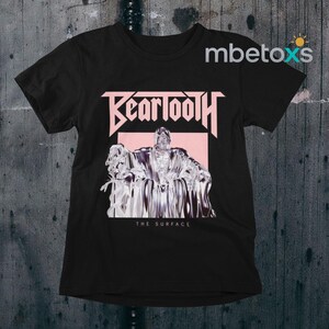 May include: Black t-shirt featuring the band name "Beartooth" in a pink, stylized font above a graphic of a figure. Below the graphic is the text "THE SURFACE". The shirt has short sleeves and a crew neck.
