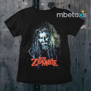 May include: Black t-shirt featuring a portrait of Rob Zombie with long hair and a beard, with green eyes and a red cross on his forehead. The name "ROB ZOMBIE" is printed in orange below the portrait.