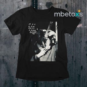 May include: Black t-shirt featuring a grayscale image of a person playing a guitar. The text "MAC DEMARCO SALAD DAYS" is printed in white on the upper left. The shirt has a crew neck and short sleeves.
