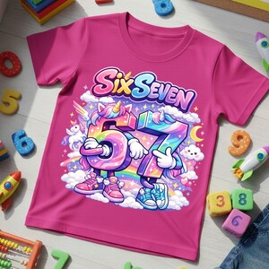 May include: A magenta t-shirt featuring the text "Six Seven" above a cartoon illustration of the numbers 6 and 7 as unicorns. The numbers are rainbow-colored and wearing sneakers. The background includes clouds, a rainbow, and a crescent moon.