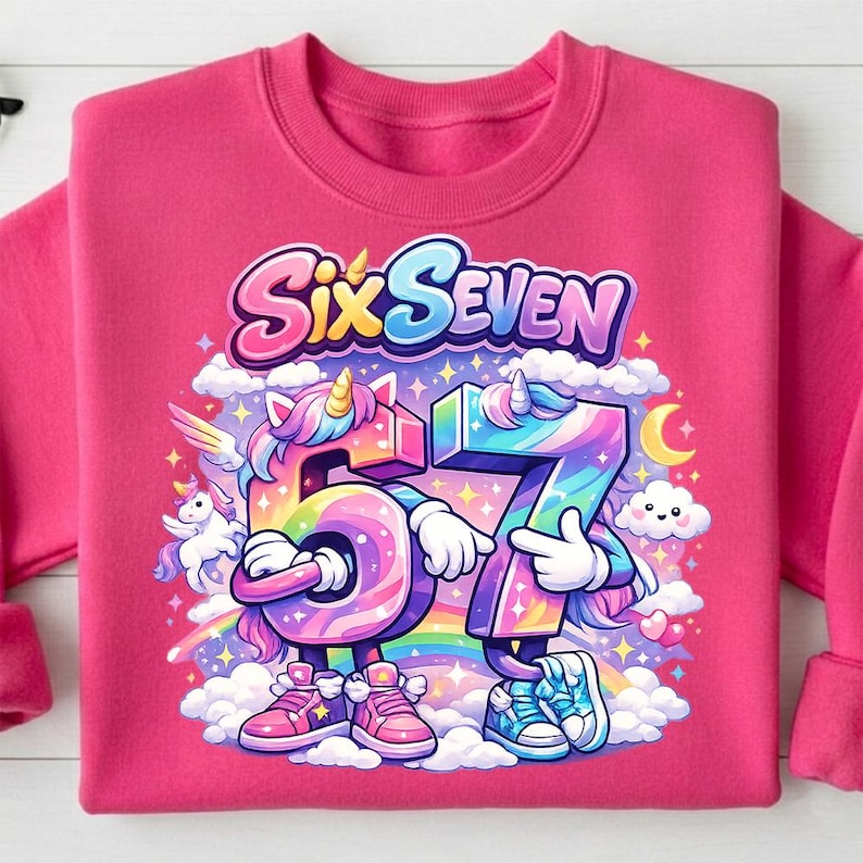 May include: A pink sweatshirt featuring a colorful graphic with the text "Six Seven". The graphic includes the numbers 5 and 7 as cartoon unicorns, a rainbow, clouds, and a crescent moon.