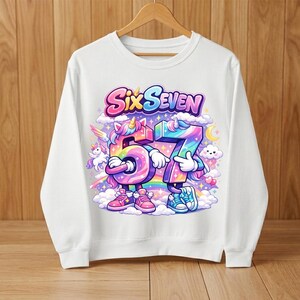 May include: White sweatshirt with a colorful graphic. The graphic features the words "Six Seven" above the numbers 5 and 7, which are designed as cartoon unicorns. The design includes a rainbow, clouds, and stars.
