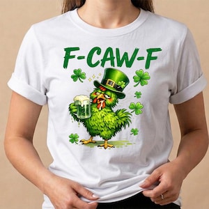 Op de afbeelding: Wit T-shirt met een groene afbeelding van een kip met een groene hoge hoed en een bierpul. De tekst "F-CAW-F" staat boven de kip, met klavers rond het ontwerp. Een leuk shirt met St. Patrick's Day thema.