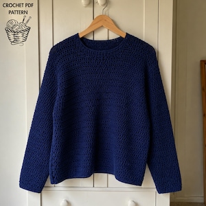 Può includere: Un maglione blu scuro a maniche lunghe con scollo rotondo, realizzato con un motivo a uncinetto testurizzato. Il capo è appeso a una gruccia di legno. Il testo CROCHET PDF PATTERN e un grafico di un cestino di lana sono visibili nell'angolo.