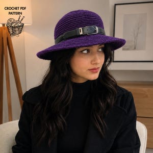 May include: A deep purple crochet hat featuring a textured, ribbed stitch pattern and a wide brim. A black leather-like belt with a silver-toned metal buckle is wrapped around the base of the crown. The text CROCHET PDF PATTERN is visible.