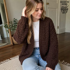 May include: A dark brown, long-sleeved, open-front cardigan with a textured, bobble-like stitch pattern, ribbed cuffs, and a ribbed collar. The garment is worn over a plain white t-shirt and light blue jeans. Text in the corner reads CROCHET PDF PATTERN.