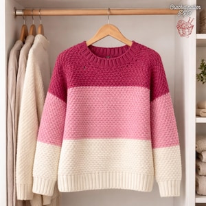 May include: A colour-blocked sweater in shades of pink and cream hangs on a wooden hanger. The sweater features a textured knit design and a ribbed neckline and cuffs. The top section is a deep pink, followed by a lighter pink band, and a cream-coloured bottom.