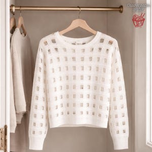 Könnte beinhalten: Ein weißer Pullover mit offenem Gewebe und gitterartigem Muster hängt an einem Holzbügel in einem Schrank. Das langärmlige Oberteil hat einen Rundhalsausschnitt und gerippte Bündchen. Das Bild enthält auch ein Logo mit der Aufschrift "Crochet pattern PDF".