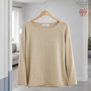 Puede incluir: Un suéter beige de manga larga con cuello redondo cuelga de una percha de madera. El suéter tiene un corte holgado y un diseño sencillo. El texto "Maantiek" es visible en el escote. La imagen también incluye un pequeño gráfico con el texto "Crochet pattern PDF".